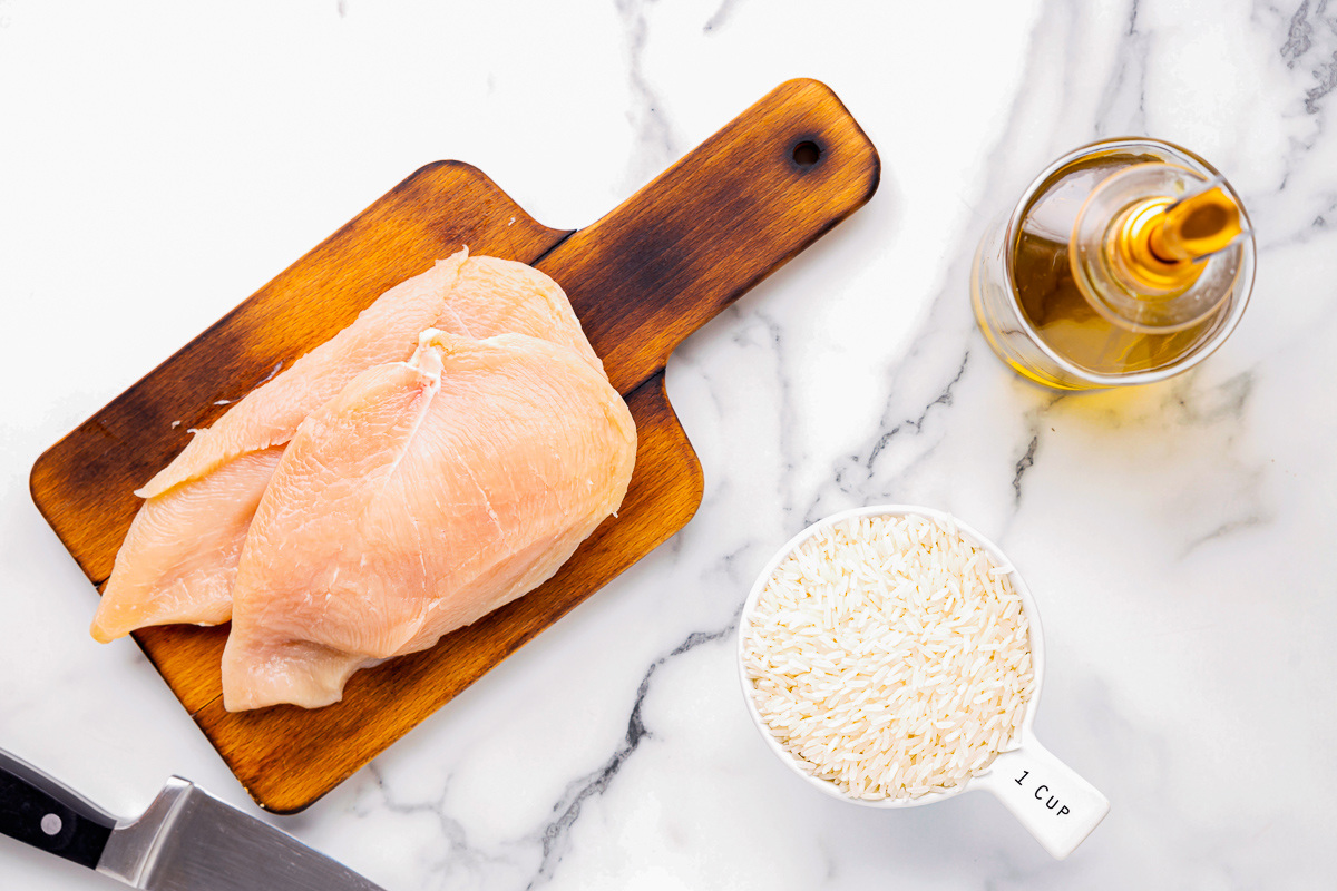 a raw chicken breast sliced in half on a small wooden cutting board next to spices and rice.