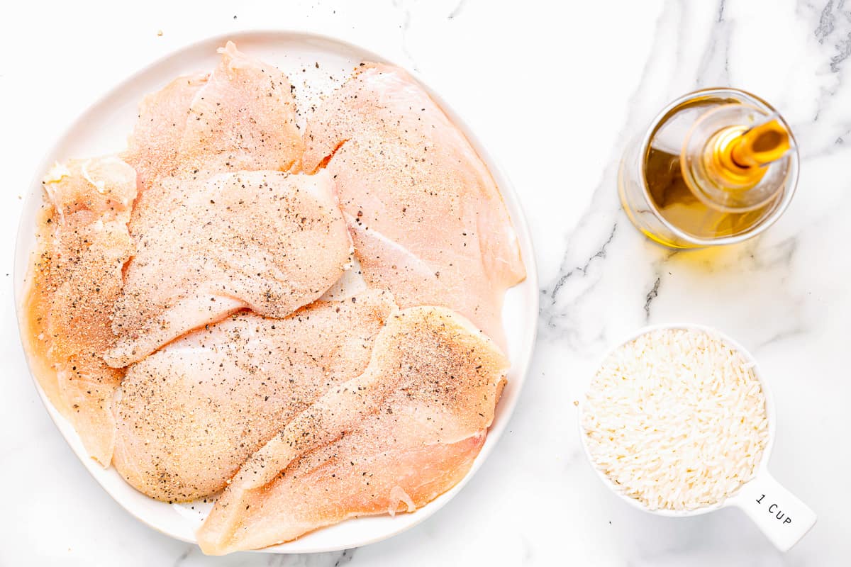 raw chicken cutlets on a white plate next to spices and rice.