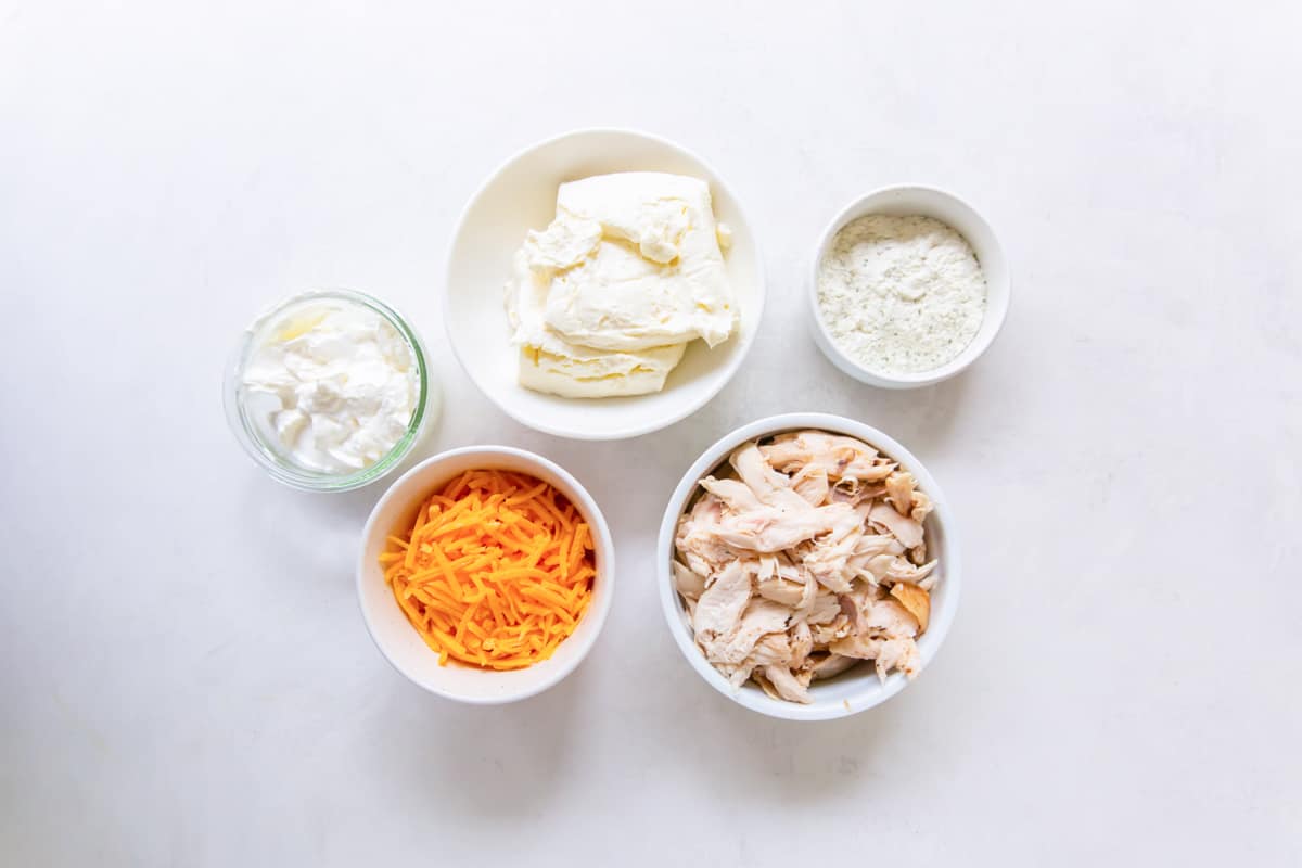 5 ingredients for ranch chicken dip arranged in small dishes: ranch seasoning, shredded cheese, cream cheese, sour cream, and shredded chicken.