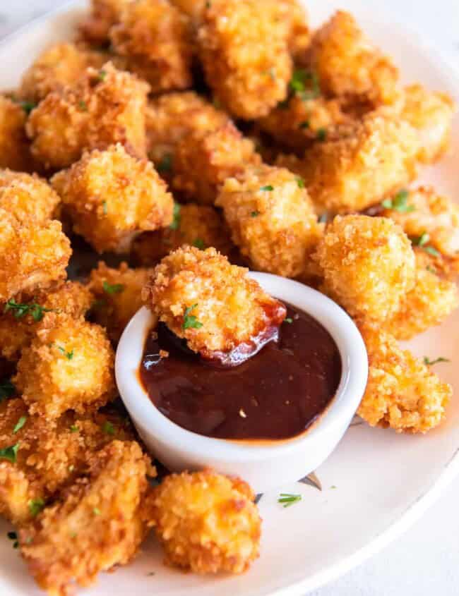 a piece of popcorn chicken being dipped into dipping sauce on a platter of popcorn chicken.