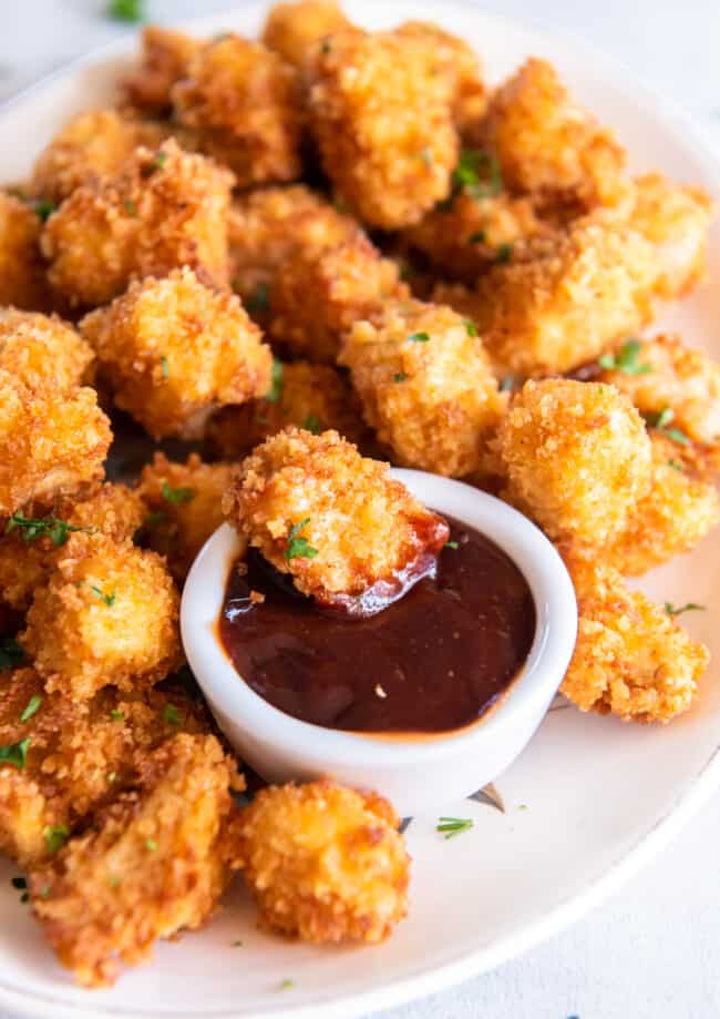 a piece of popcorn chicken being dipped into dipping sauce on a platter of popcorn chicken.