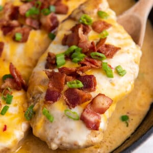 a wooden spoon lifting a cheesy bacon ranch chicken breast from a cast iron skillet.