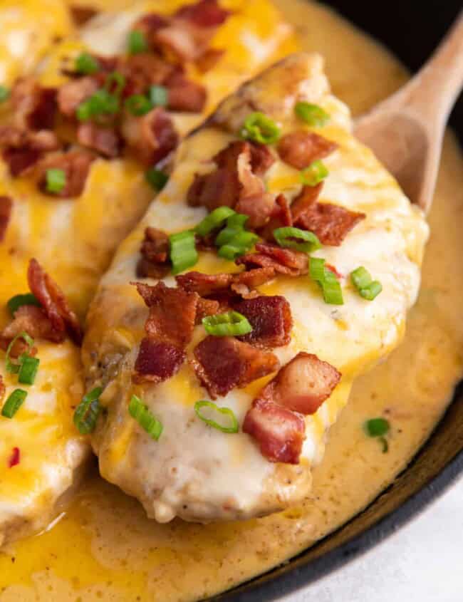 a wooden spoon lifting a cheesy bacon ranch chicken breast from a cast iron skillet.