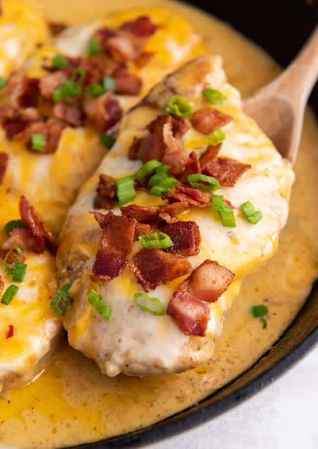 a wooden spoon lifting a cheesy bacon ranch chicken breast from a cast iron skillet.