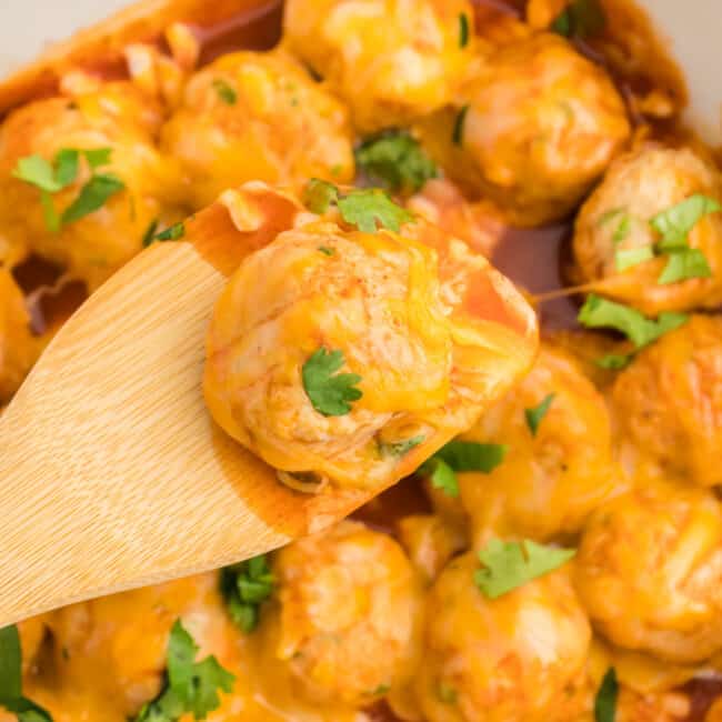 close up of a wooden spoon lifting a chicken enchilada meatball from a white oval baking dish.