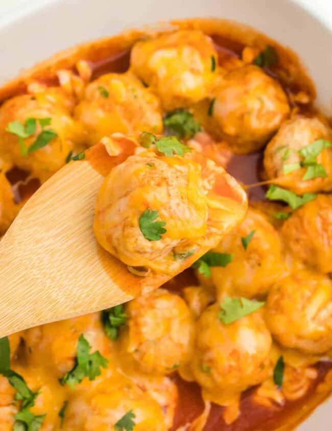 close up of a wooden spoon lifting a chicken enchilada meatball from a white oval baking dish.