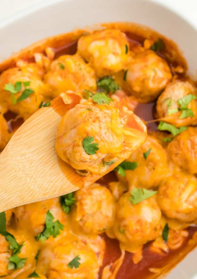 close up of a wooden spoon lifting a chicken enchilada meatball from a white oval baking dish.