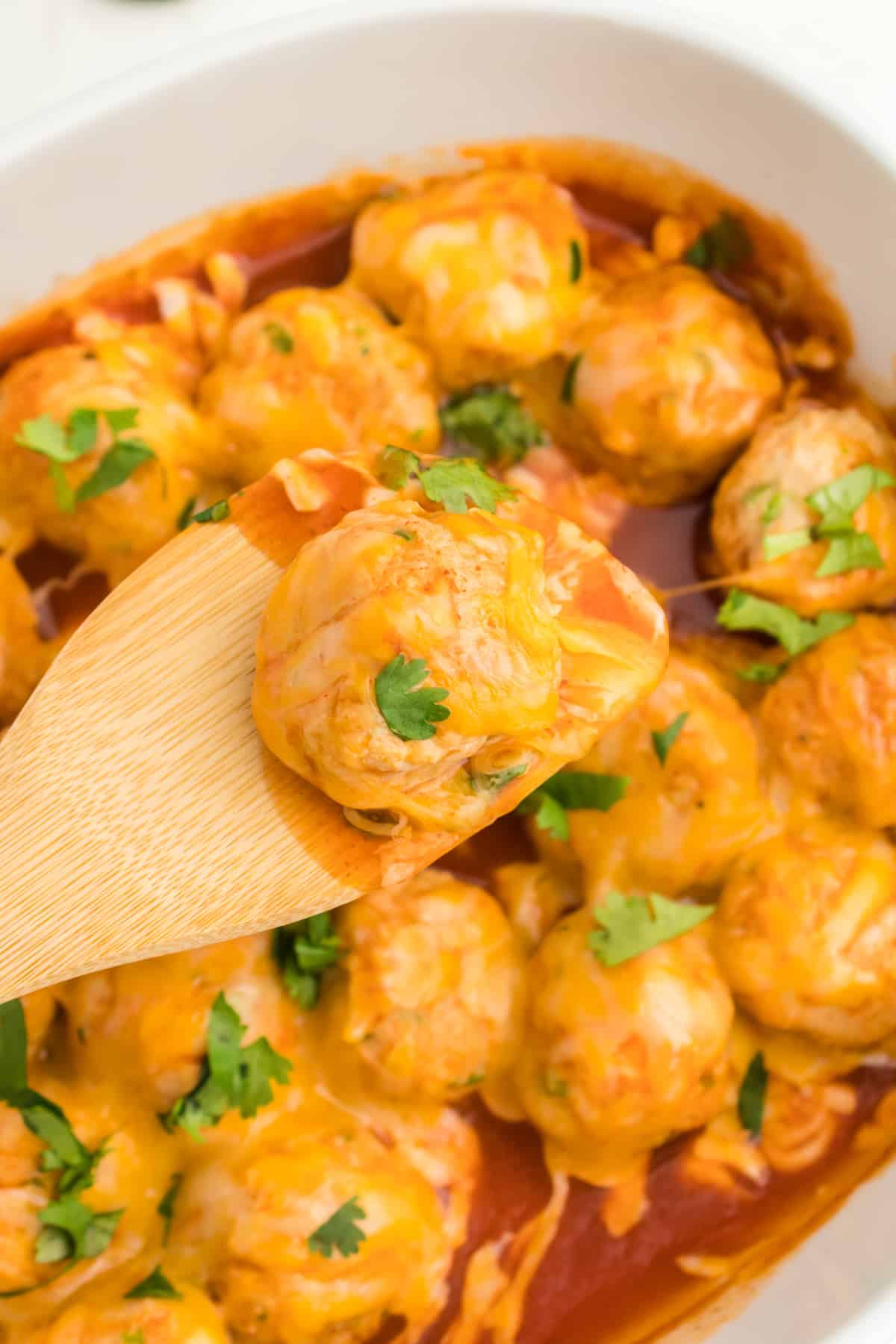 close up of a wooden spoon lifting a chicken enchilada meatball from a white oval baking dish.