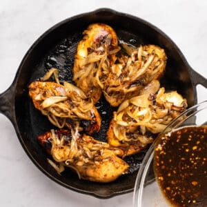 balsamic marinade poured over 5 cooked balsamic glazed chicken breasts with caramelized onions in a cast iron pan.