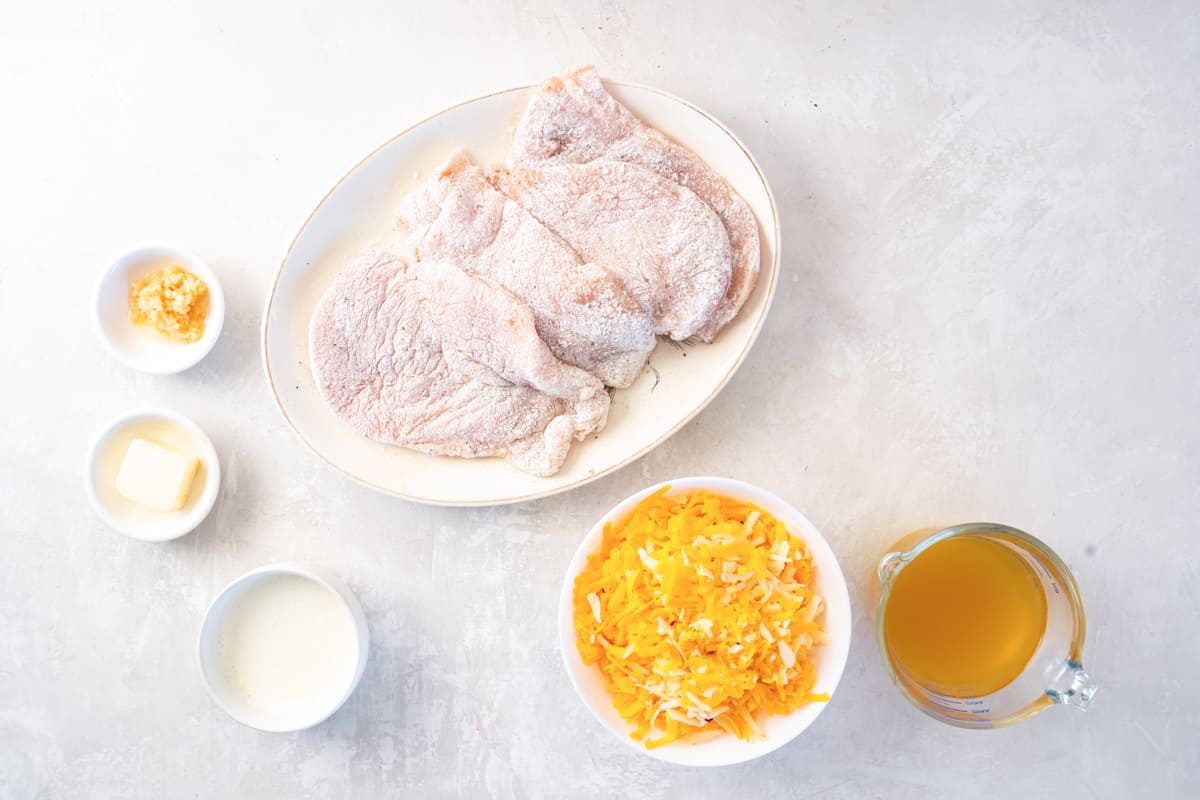 seasoned and flour coated chicken breasts on a white plate next to remaining ingredients for cheesy bacon ranch chicken.