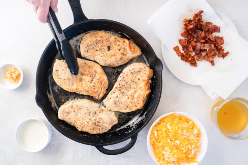 4 chicken breasts cooking in a cast iron skillet; a pair of tongs is flipping one breast.