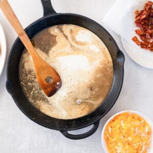 cream added to cheesy bacon ranch chicken sauce in a cast iron skillet with a wooden spatula.