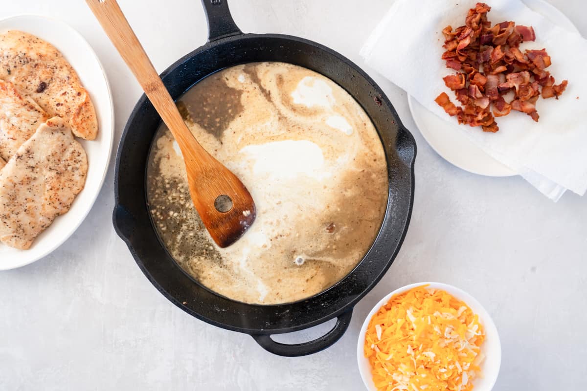 cream added to cheesy bacon ranch chicken sauce in a cast iron skillet with a wooden spatula.