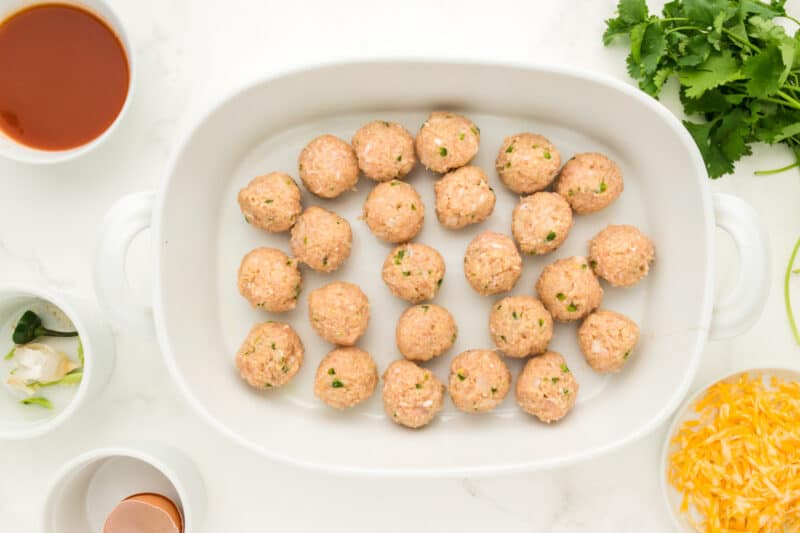 24 raw chicken enchilada meatballs in a white oval baking dish.