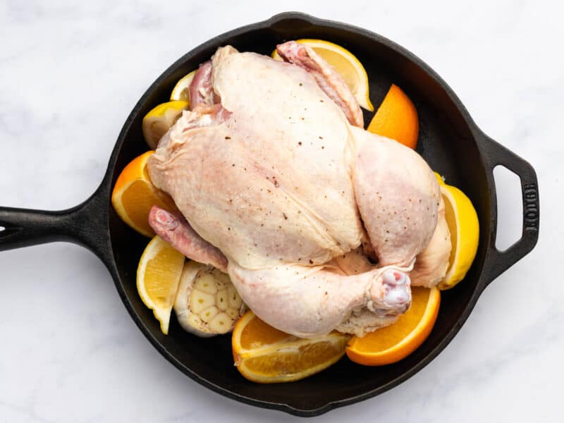 a raw whole chicken on a bed of garlic and cut citrus in a cast iron skillet.