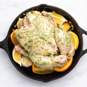 a whole raw chicken coated in herb butter on a bed of whole garlic and citrus pieces in a cast iron skillet.