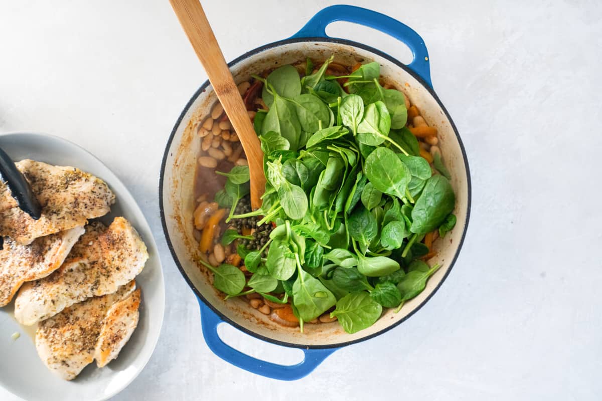 spinach added to italian chicken skillet in a cast iron pan with a wooden spoon.