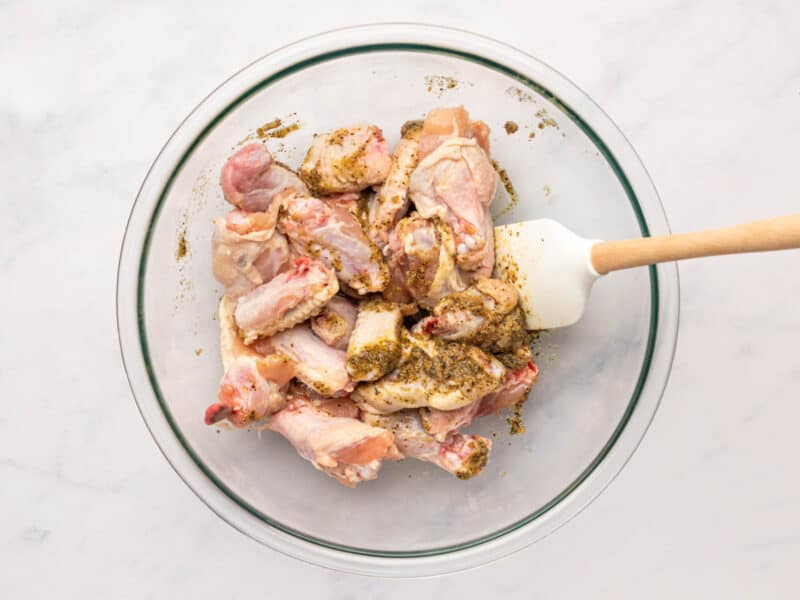 raw chicken wings tossed in lemon pepper sauce in a glass bowl with a rubber spatula.