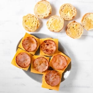 overhead view of 6 english muffins topped with cheese, an egg round, and a slice of ham.