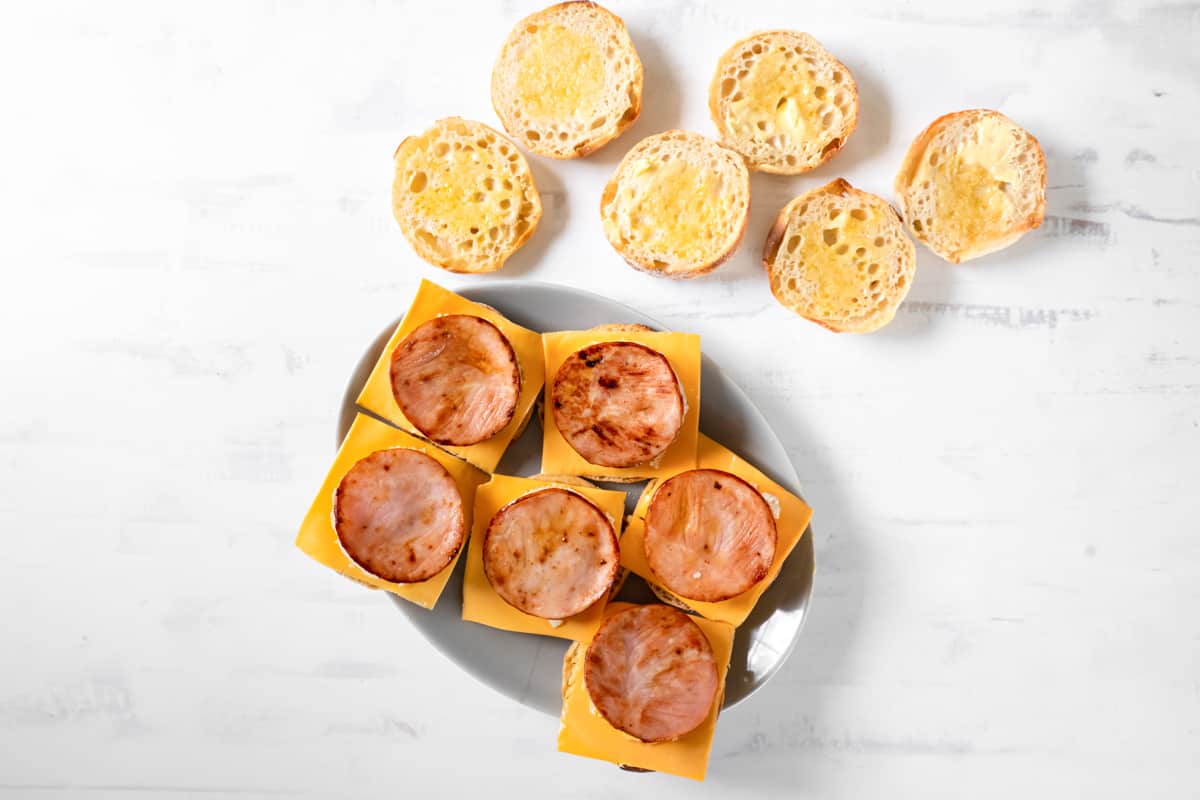 overhead view of 6 english muffins topped with cheese, an egg round, and a slice of ham.