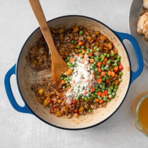 a pan with chicken, peas and carrots next to a wooden spoon.