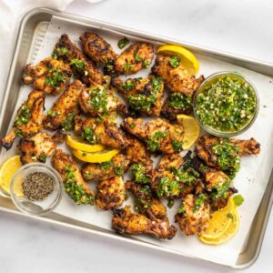 overhead view of lemon pepper wings on a baking sheet with gremolata and lemon wedges.