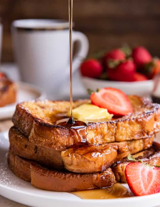syrup pouring over a 3 stack of french toast on a white plate with butter and a strawberry.