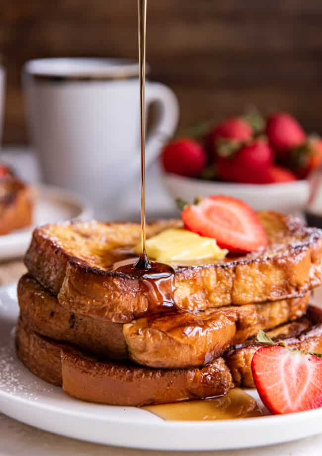 syrup pouring over a 3 stack of french toast on a white plate with butter and a strawberry.