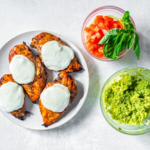 4 grilled chicken breasts on a white plate with cheese on top next to diced tomatoes and guacamole in glass bowls.