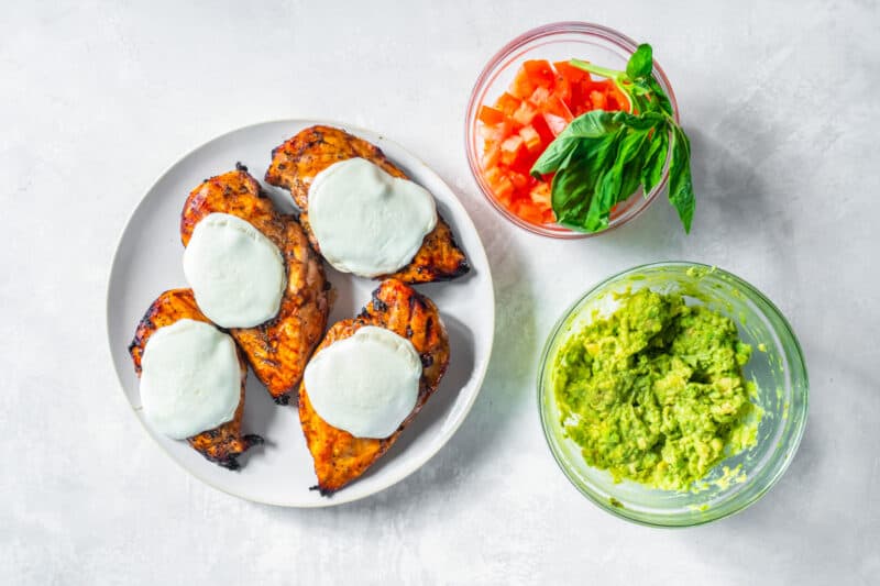 4 grilled chicken breasts on a white plate with cheese on top next to diced tomatoes and guacamole in glass bowls.
