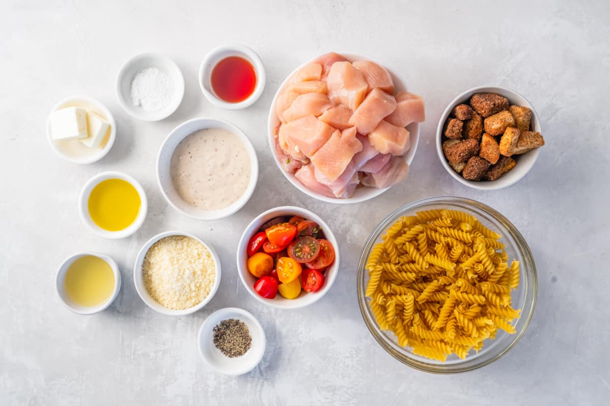 Ingredients for chicken caesar pasta salad arranged in dishes: cubes of chicken, croutons, pasta, tomatoes, caesar dressing, parmesan.