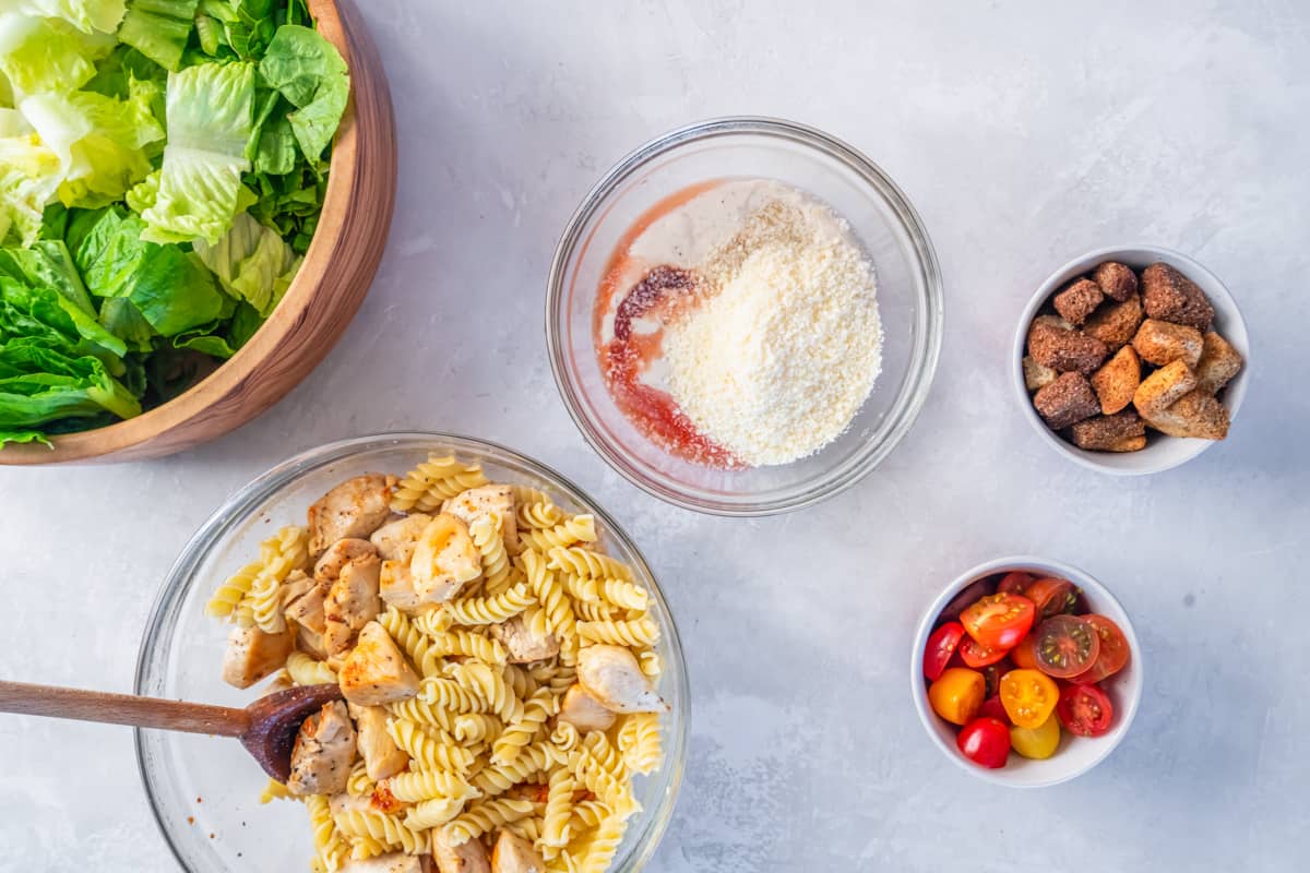 Dressing ingredients in a glass bowl; next to bowls of pasta and chicken, croutons, lettuce, and tomatoes.