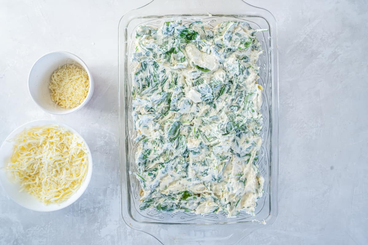 spinach dip mixture spread over chicken and artichokes in a glass casserole dish.