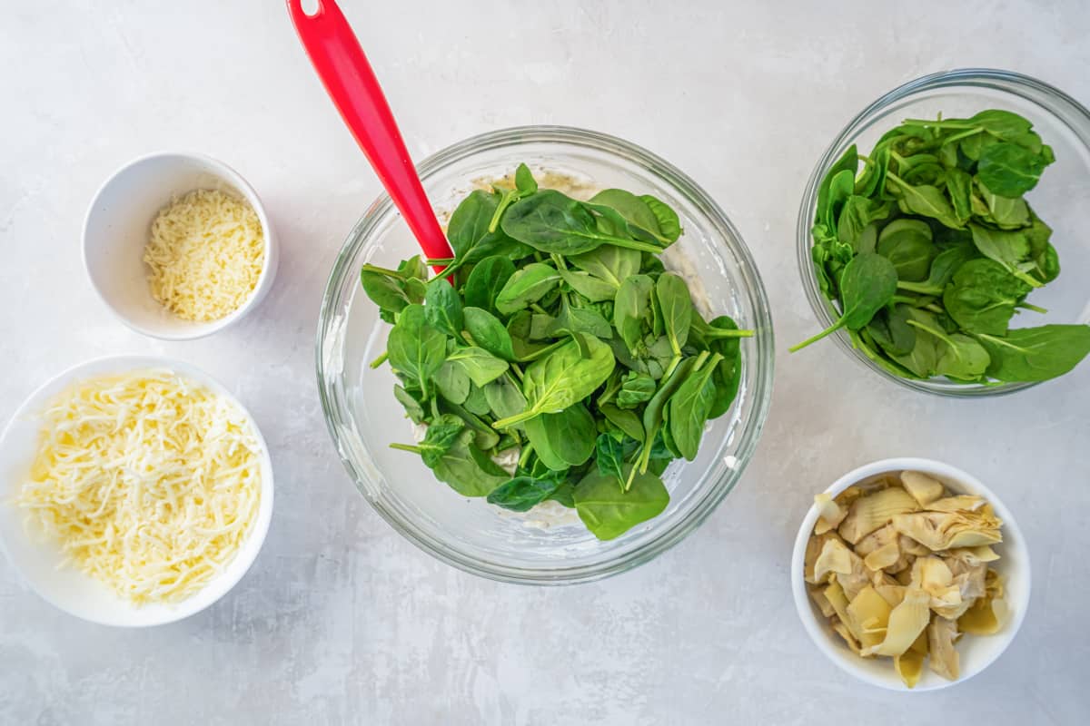 spinach added to cream cheese mixture in a glass bowl with a red rubber spatula.