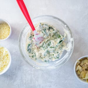 spinach dip mixture in a glass bowl with a red rubber spatula.
