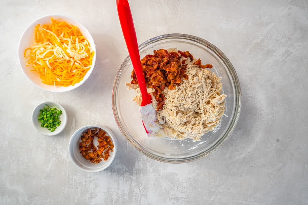 chicken and bacon added to sour cream and cheese mixture in a glass bowl with a rubber spatula.