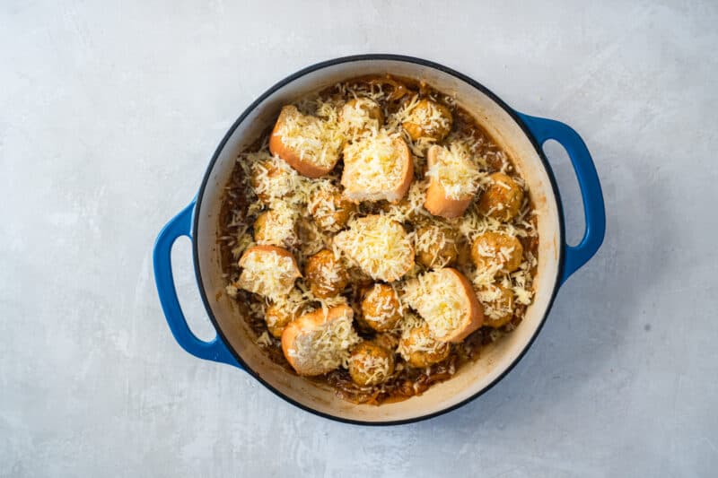 bread and cheese on top of french onion meatballs in a blue and white dutch oven.