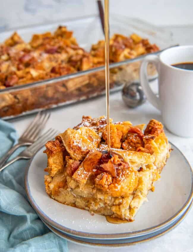 three-quarters view of maple syrup drizzled over a slice of chicken and waffle casserole on a white plate.