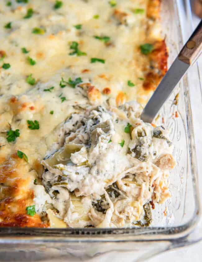 three-quarters view of a spoon lifting a scoop of spinach artichoke chicken casserole from a glass baking dish.