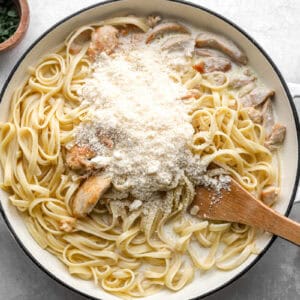 grated cheese over chicken alfredo in a pan with a wooden spatula.