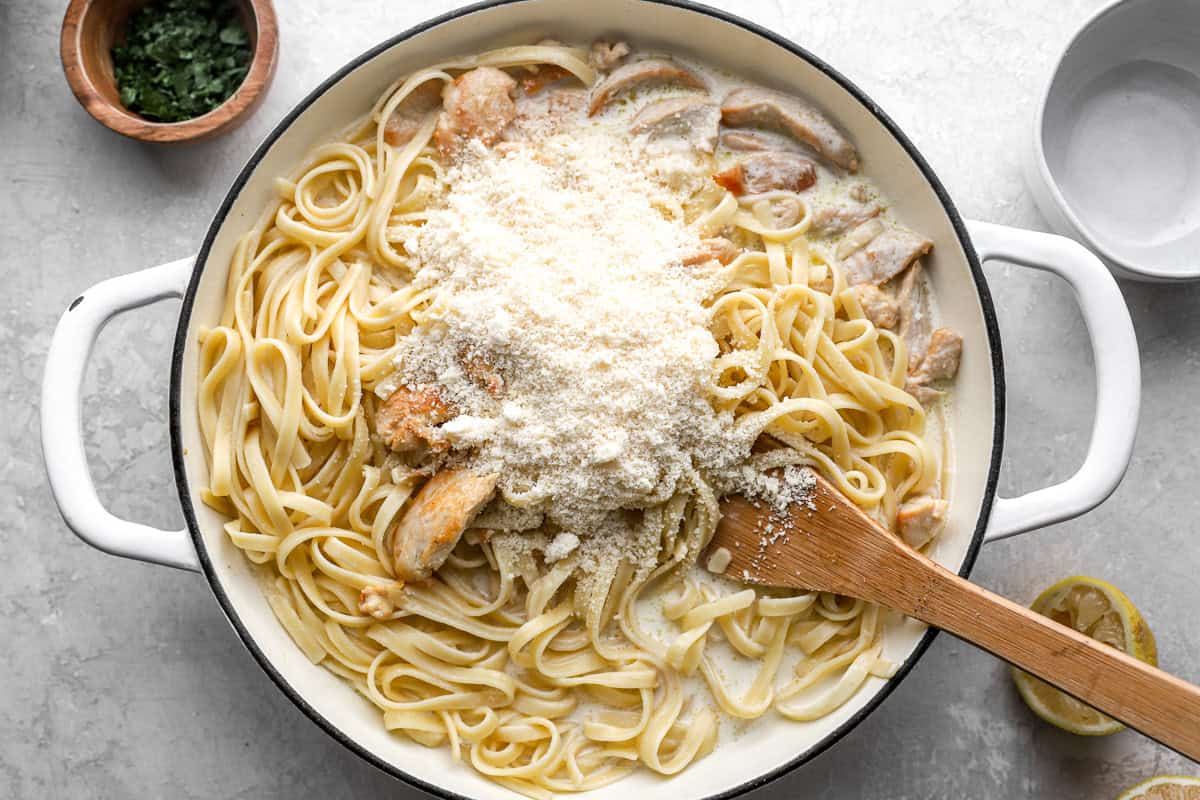 grated cheese over chicken alfredo in a pan with a wooden spatula.