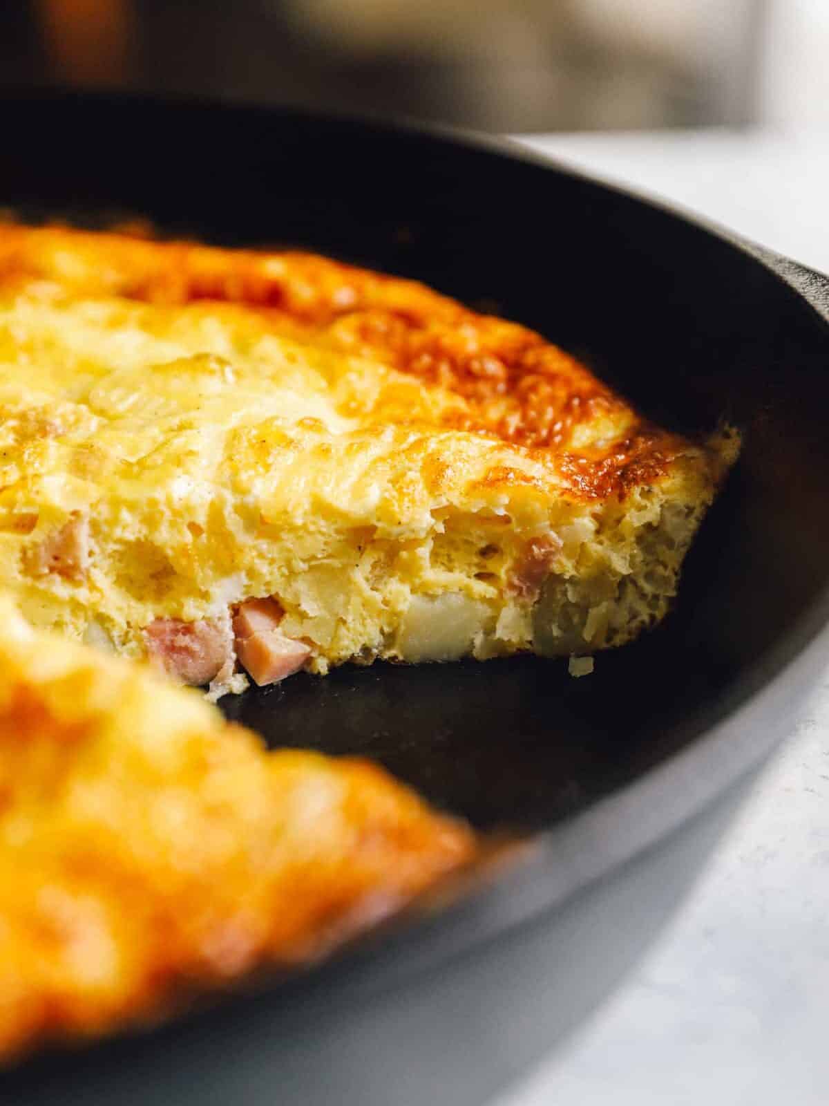 close-up three-quarters view of a ham and cheese frittata in a cast iron pan missing a slice.