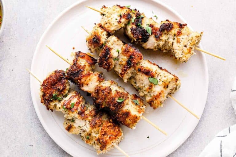 A white plate holds four grilled chicken kebabs on wooden skewers. The chicken pieces are cooked with a golden-brown exterior and sprinkled with herbs, giving them a slightly charred look. The presentation is clean, with no additional garnishes visible.