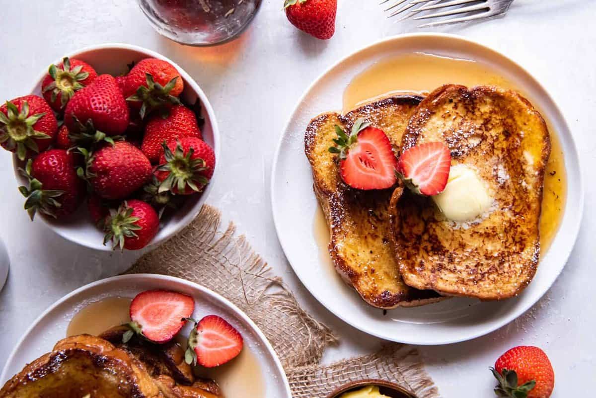overhead view of slices of french toast on a white plate with syrup, butter, and a strawberry.
