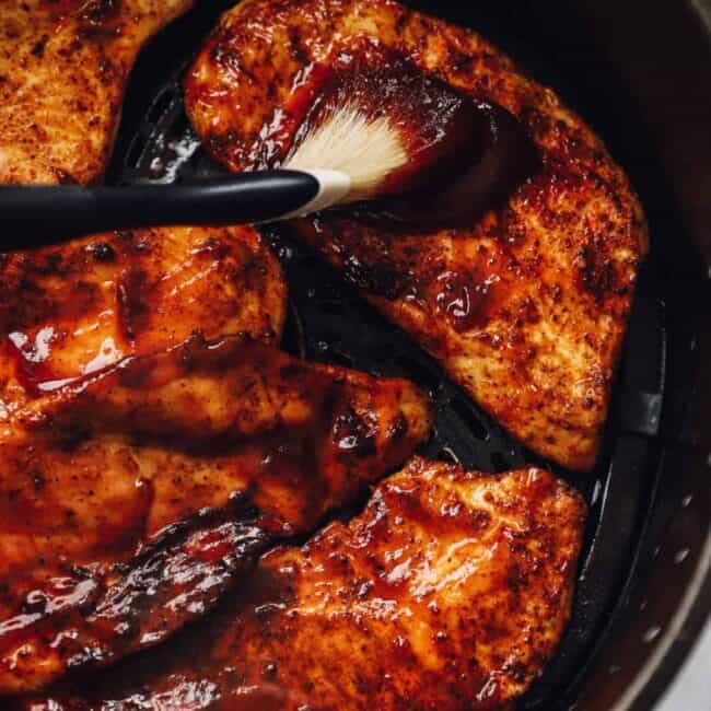 brushing BBQ sauce onto cooked chicken breasts in the air fryer basket