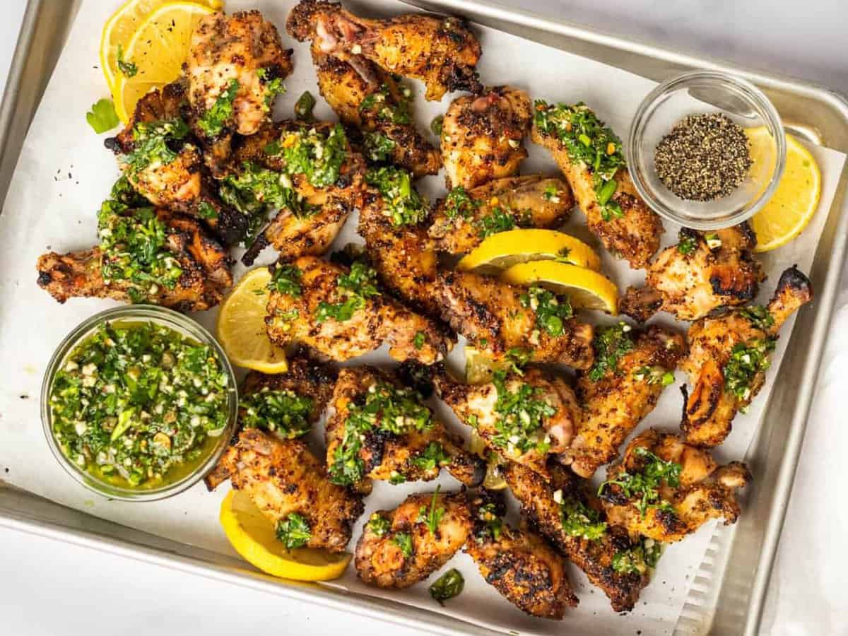 overhead view of lemon pepper wings on a baking sheet with gremolata and lemon wedges.