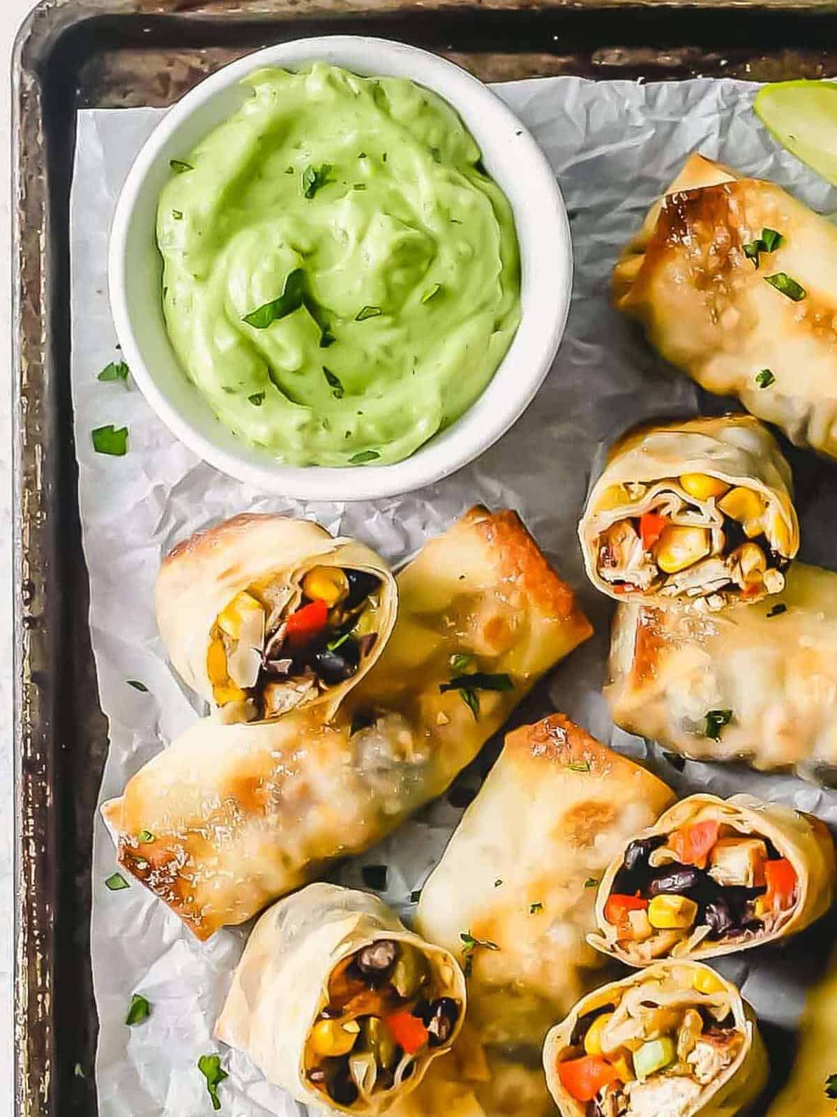 southwest chicken egg rolls on a sheet pan with a bowl of avocado dip