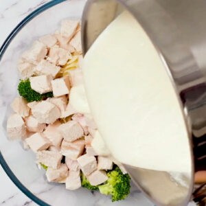 pouring cheese sauce over broccoli, rotini, and chicken in a glass bowl.