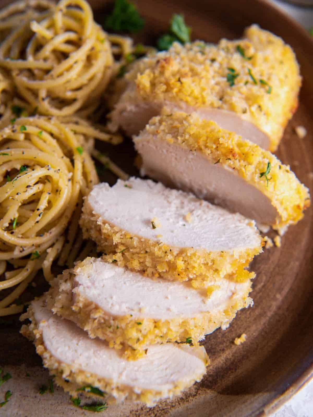 close up of a sliced parmesan ranch chicken breast on a brown plate with nests of pasta.