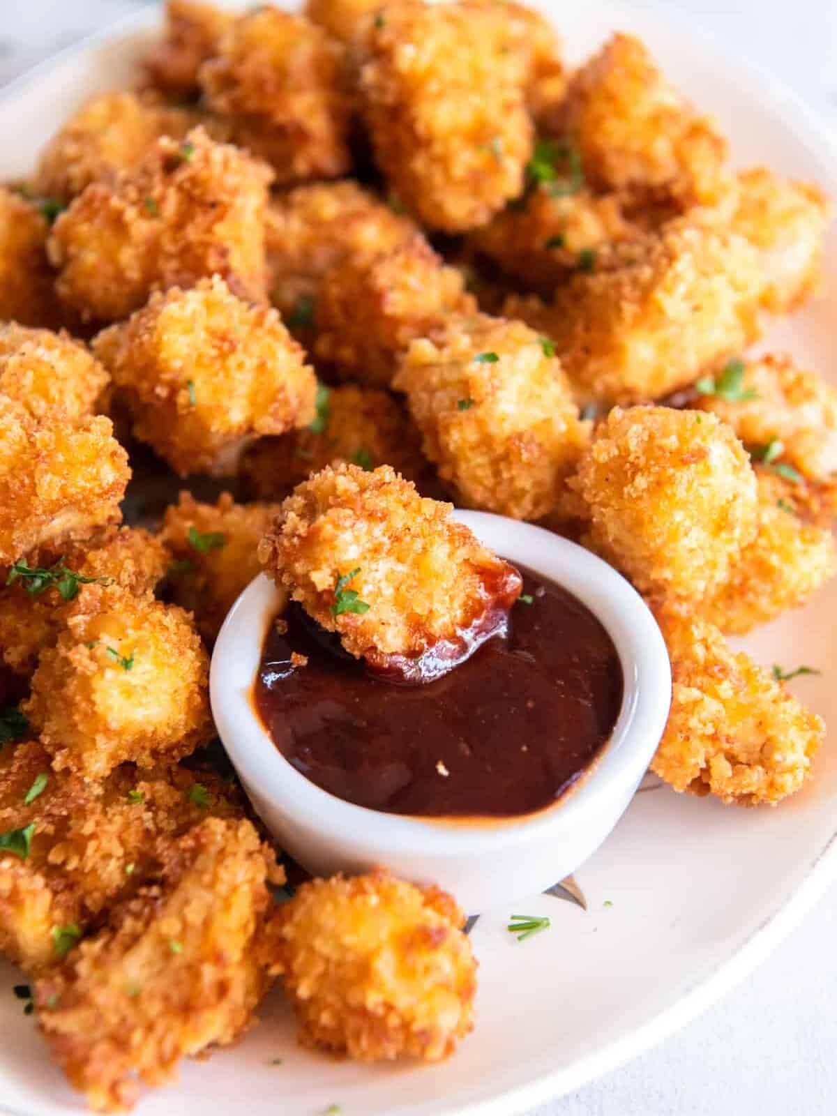 a piece of popcorn chicken being dipped into dipping sauce on a platter of popcorn chicken.